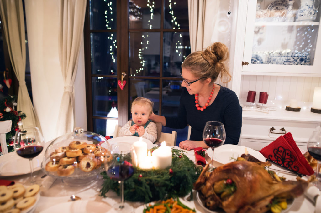 Beautiful young mother with her cute little son celebrating Christmas together with their family. Food and Christmas wreath with candles laid on a table.